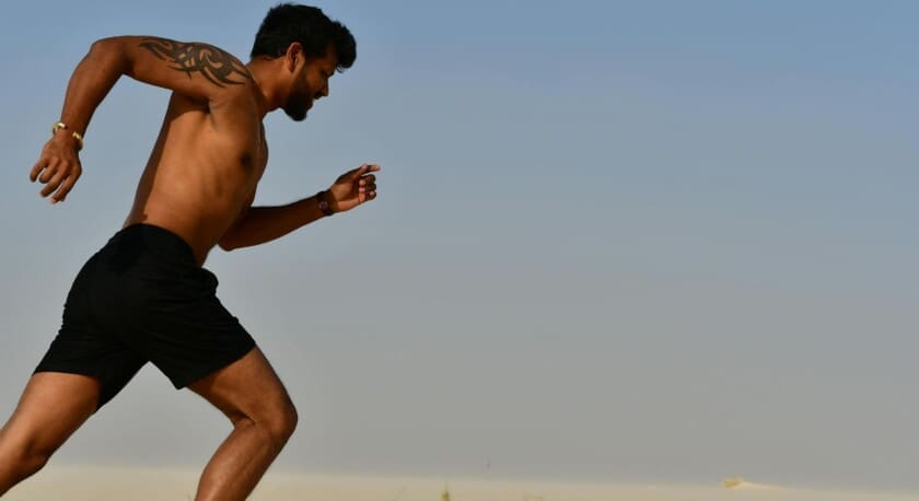 A man is running in a desert landscape, wearing black shorts and no shirt. He has a tattoo on his upper arm and appears to be in mid-stride, showcasing a dynamic running posture. The background features a clear blue sky and light-colored sand, emphasizing the vastness of the desert.