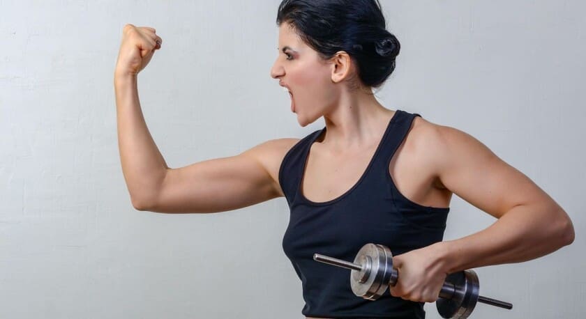 A woman is flexing her bicep while holding a dumbbell in her other hand. She has a determined expression, with her mouth open as if shouting or cheering. She is wearing a black tank top, and her hair is styled in a bun. The background is a light, neutral color, enhancing the focus on her strong pose.