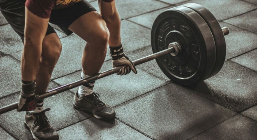 A person is preparing to lift a barbell with heavy weights on each side. The individual is wearing a maroon shirt and black shorts, with gloves on their hands for grip. The barbell rests on a textured rubber gym floor, and the person's feet are positioned firmly on the ground, wearing athletic shoes. The scene conveys a focused moment in a weightlifting session.