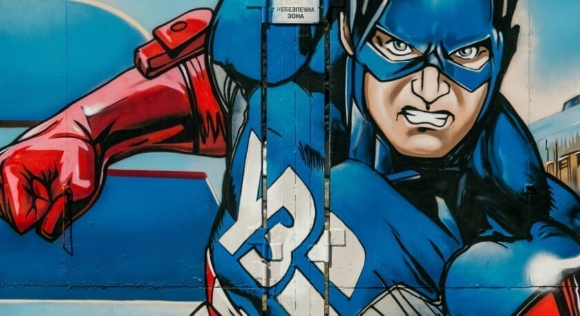 A vibrant mural features a muscular superhero in a blue and red costume, striking a dynamic pose as if charging forward. The character has a fierce expression, with a blue mask and a prominent emblem on the chest. The background is a gradient of blue, enhancing the action-packed feel of the artwork. A sign in the upper part of the mural reads a cautionary message in a different language.