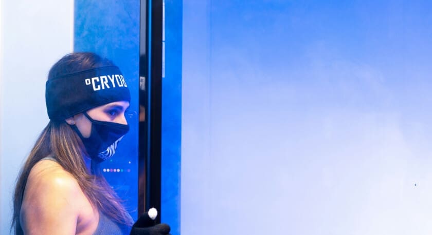 A woman stands beside a cryotherapy chamber, wearing a black headband and a matching mask with the word "CRYO" on it. She has long hair and is dressed in a sleeveless top. Her gloved hand is positioned near the chamber, which emits a misty vapor, creating a cool blue ambiance in the environment. The background features a gradient of blue tones, enhancing the chilly atmosphere.