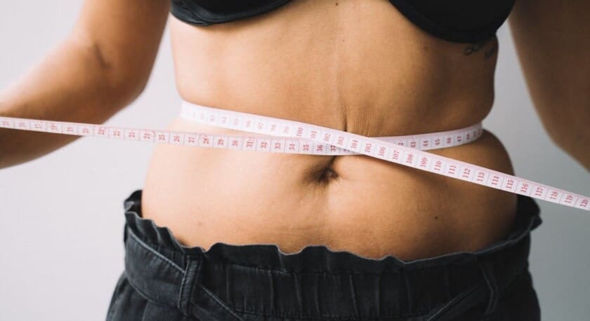 A close-up view of a person's midsection, showing a bare abdomen with a measuring tape wrapped around the waist. The individual is wearing a black bra and dark pants. The measuring tape is marked with measurements in centimeters. The background is a neutral color, emphasizing the focus on the waist measurement.
