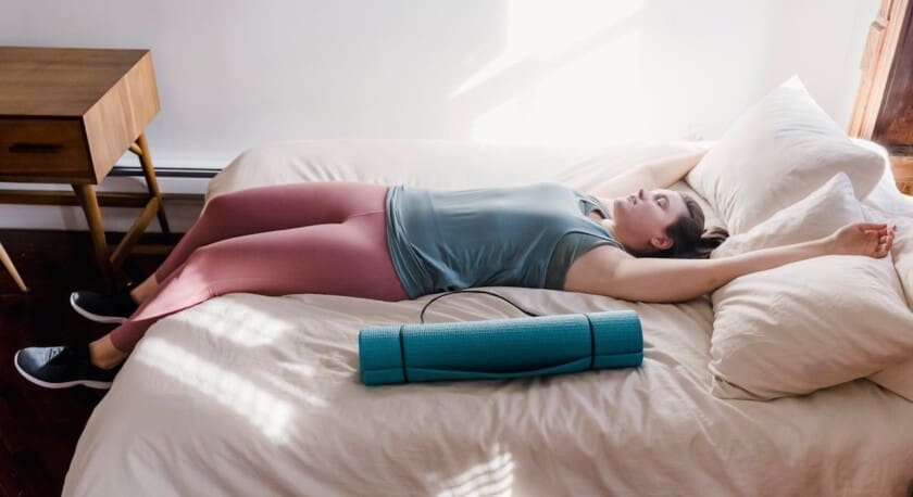 A person is lying on a bed with their arms extended above their head and legs stretched out. They are wearing a light blue tank top and pink leggings. A yoga mat in a teal color is placed beside them on the bed. The room is well-lit with natural light coming through a window, casting soft shadows. A wooden nightstand is visible next to the bed.