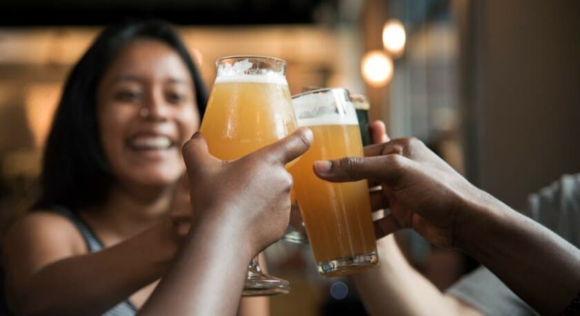 A group of hands is raised in a toast, holding glasses filled with a light-colored beverage, likely beer. In the background, a smiling woman is enjoying the moment, with soft lighting creating a warm atmosphere. The glasses have a frothy top, and the setting appears to be a casual gathering, possibly in a bar or restaurant.