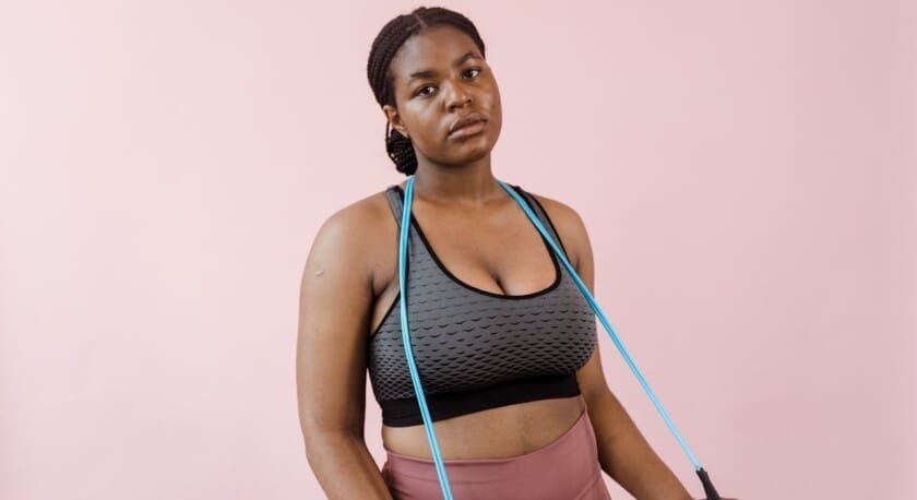 A young woman stands against a soft pink background, wearing a fitted gray sports bra with a textured pattern and high-waisted pink leggings. She holds a pair of blue resistance bands, draped around her shoulders. Her hair is styled in braids, and she has a serious expression, suggesting focus and determination.