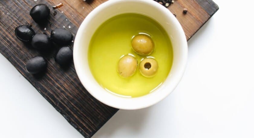 A small white bowl filled with green olive oil contains three green olives. Surrounding the bowl are several black olives and a scattering of black peppercorns on a wooden cutting board. The background is a light surface, enhancing the colors of the olives and oil.