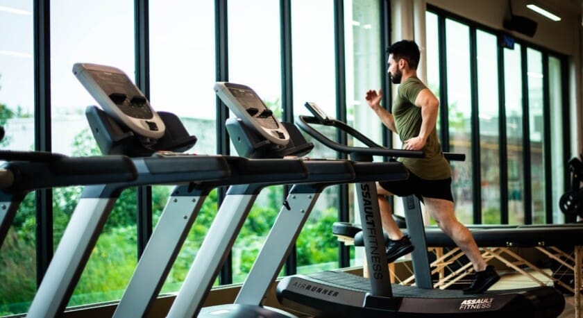 A man is running on a treadmill in a gym setting, wearing a green shirt and black shorts. The treadmill is part of a row of fitness equipment, with large windows in the background allowing natural light to fill the space. Outside the windows, greenery is visible, creating a refreshing atmosphere. The gym appears modern and well-lit.