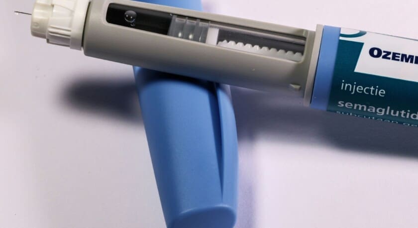 A gray and blue injection pen is positioned on a light surface. The pen features a clear window showing a small bubble of liquid inside, and it has a needle at the tip. Next to the pen is a blue cap, which is cylindrical in shape. The pen is labeled with the name "Ozempic" and mentions "semaglutide" and "injectie.