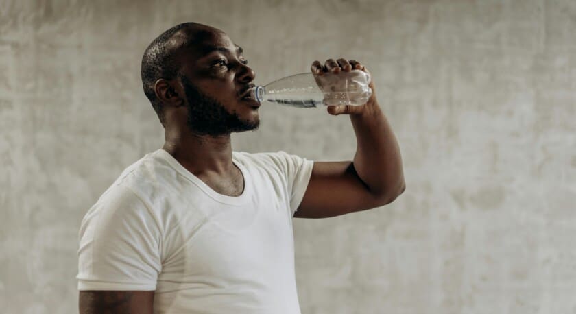 A man is drinking from a clear plastic water bottle. He is wearing a plain white t-shirt and has a short beard. The background features a textured, light-colored wall, creating a neutral setting. The focus is on the man as he tilts the bottle towards his mouth, appearing to hydrate.
