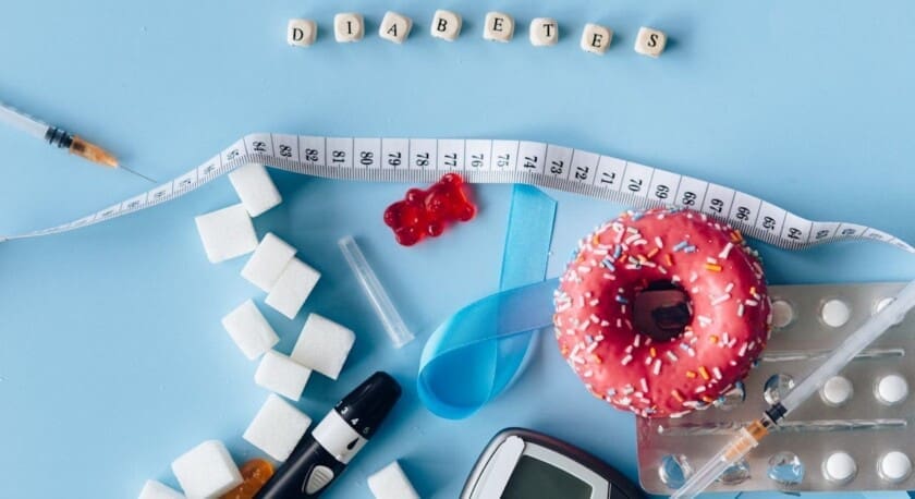 A flat lay arrangement features various items related to diabetes management. A pink frosted donut with colorful sprinkles sits prominently. Nearby, there are several sugar cubes, a blue awareness ribbon, and a glucose meter. A syringe and a vial are also present, alongside a strip of medication tablets. The word "DIABETES" is spelled out with wooden letter blocks, and a measuring tape is partially coiled in the scene. The background is a light blue, creating a vibrant contrast with the items.