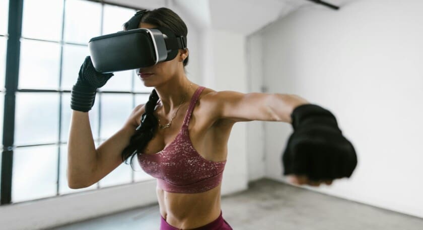 A woman is engaged in a virtual reality workout, wearing a VR headset and black gloves. She is in a fighting stance, with her right arm extended forward as if throwing a punch. She has a toned physique, wearing a maroon sports bra and matching high-waisted leggings. The background features large windows that allow natural light to illuminate the space, which has a minimalist, industrial feel.