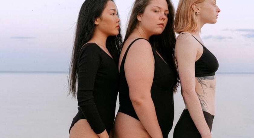 Three women stand side by side on a beach, facing slightly away from the camera. They are wearing black swimsuits, with varying styles and fits. The woman on the left has long, dark hair, the woman in the center has medium-length, wavy hair, and the woman on the right has short, light-colored hair. The background features a calm sea and a pastel sky, suggesting a serene atmosphere.