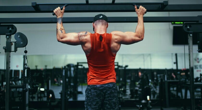 A person is performing a pull-up on a bar in a gym setting. They are wearing a bright orange sleeveless shirt and camouflaged shorts. The focus is on their muscular back and arms, showcasing significant muscle definition. The gym is equipped with various workout machines and weights in the background, and there is a mirror reflecting part of the gym environment.