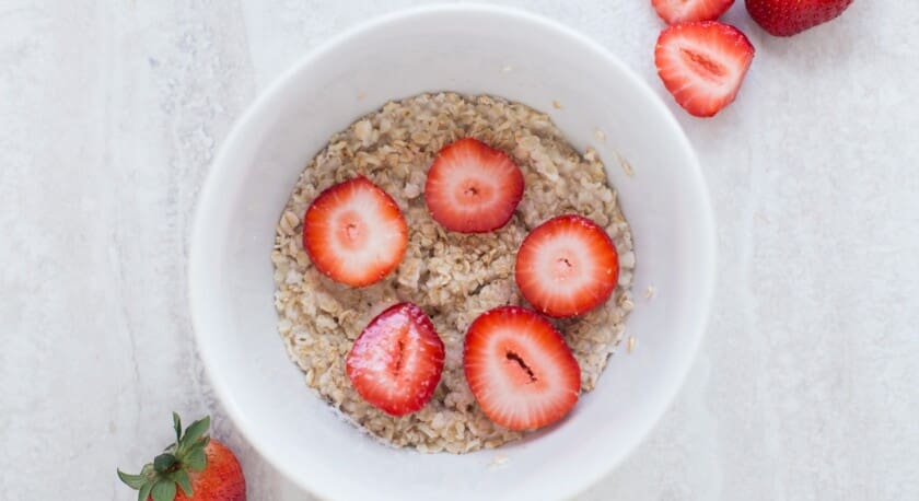 A white bowl filled with oatmeal is topped with several slices of fresh strawberries. The oatmeal has a creamy texture, and the bright red strawberries contrast beautifully against it. To the side of the bowl, there are two whole strawberries, one upright and one cut in half, showcasing their juicy interior. The background features a light-colored, textured surface.