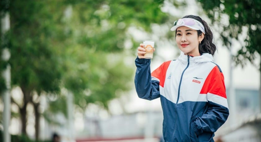 A woman stands outdoors, holding a cup of beverage with a logo. She wears a sporty jacket that features red, white, and blue colors, along with a visor. The background is filled with greenery, suggesting a park or outdoor setting. Her expression is cheerful, and she appears to be enjoying her drink.