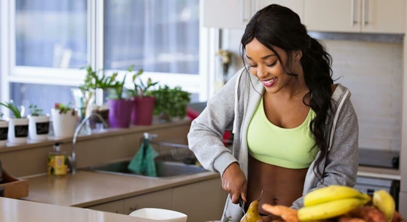A woman is preparing food in a bright kitchen. She is wearing a lime green crop top and a gray hoodie, smiling as she slices a pear. In front of her, a bowl contains strawberries, and there is a pile of sweet potatoes and bananas nearby. The kitchen features potted plants on the counter and a window that lets in natural light.