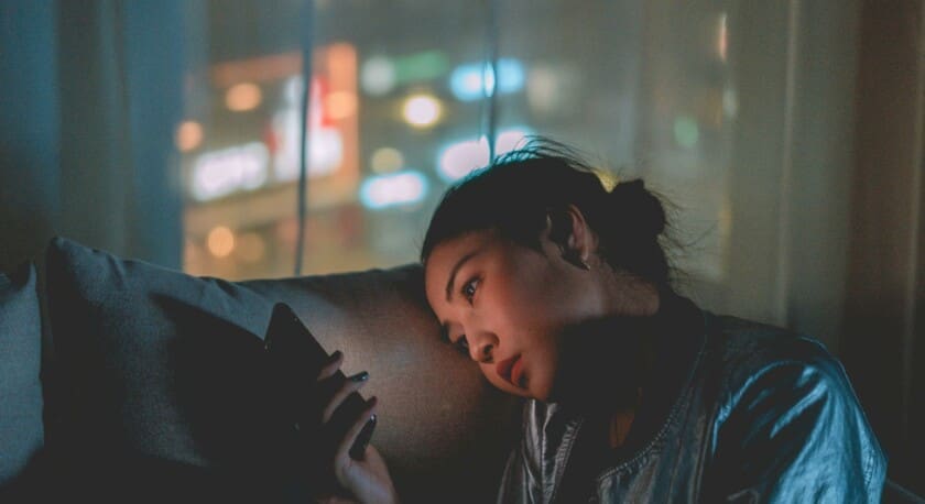 A young woman sits on a couch, leaning her head against her hand while looking at her smartphone. She has a contemplative expression, with soft lighting highlighting her features. The background shows blurred city lights through a window, creating a moody atmosphere. She wears a shiny, light-colored jacket, and her hair is pulled back in a bun. The overall scene conveys a sense of introspection and connection to the digital world.