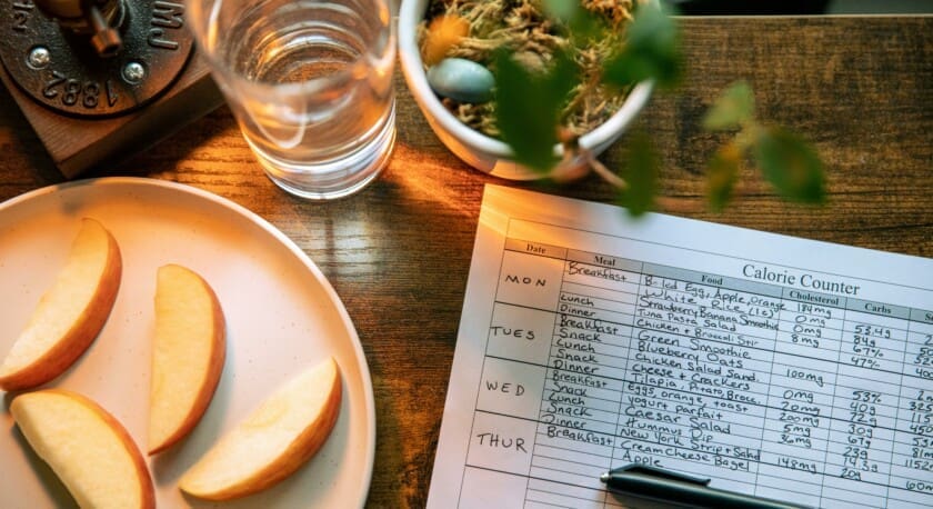 A plate with several slices of red apple is positioned on a wooden table. Nearby, a glass of water sits next to a small potted plant. A sheet of paper labeled "Calorie Counter" is partially visible, detailing meals and their nutritional information for the week. The setting is well-lit, creating a warm ambiance.