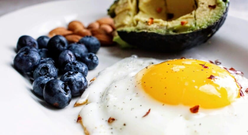 A plate features a sunny-side-up egg with a bright yellow yolk, surrounded by a sprinkle of black pepper and red chili flakes. Next to the egg, there are clusters of fresh blueberries and a handful of almonds. An avocado, cut in half, is positioned at the top right, revealing its creamy green flesh. The overall presentation is colorful and appetizing.