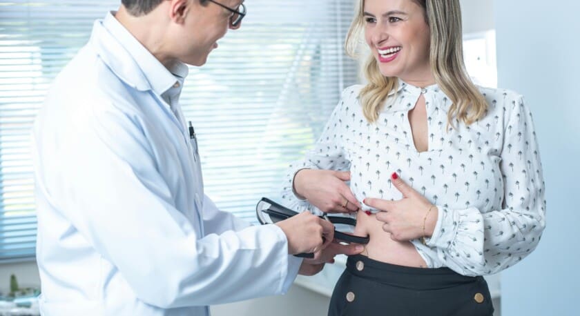 A healthcare professional in a white lab coat is examining a woman in a patterned blouse. The woman is smiling and pointing to her abdomen, while the professional is using a handheld device to assess her. The setting appears to be a medical office with natural light coming through blinds. The woman is wearing high-waisted black pants with buttons.