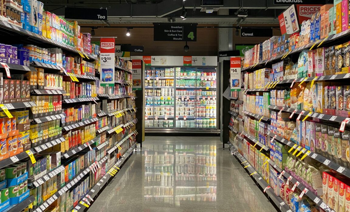 A grocery store aisle is visible, featuring shelves stocked with various beverages and breakfast items. On the left side, there are numerous colorful cartons of tea, coffee, and other drinks, with price tags indicating discounts. The right side displays a range of breakfast cereals and museli, also with promotional labels. At the end of the aisle, a refrigerated section is filled with dairy products. Overhead signs indicate the categories of items, including "Tea," "Coffee," "Breakfast," and "Cereal." The floor reflects the bright lighting from above, creating a clean and organized shopping environment.