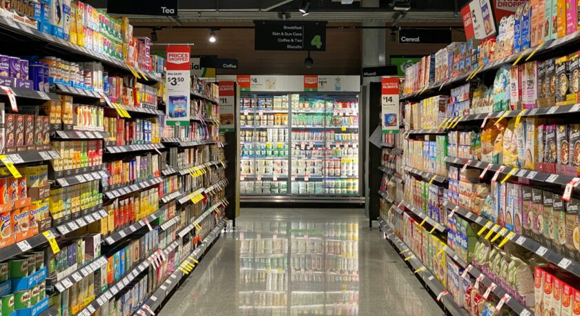 A grocery store aisle is visible, featuring shelves stocked with various beverages and breakfast items. On the left side, there are numerous colorful cartons of tea, coffee, and other drinks, with price tags indicating discounts. The right side displays a range of breakfast cereals and museli, also with promotional labels. At the end of the aisle, a refrigerated section is filled with dairy products. Overhead signs indicate the categories of items, including "Tea," "Coffee," "Breakfast," and "Cereal." The floor reflects the bright lighting from above, creating a clean and organized shopping environment.