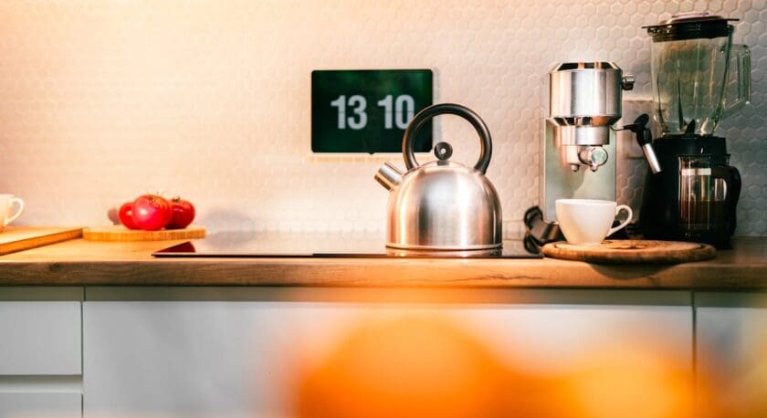 A modern kitchen countertop features a shiny silver kettle with a black handle, positioned next to a white coffee cup on a wooden cutting board. Behind the kettle, there is a sleek espresso machine and a blender. A small cluster of red tomatoes rests on a round wooden plate. In the background, a digital clock displays the time in a minimalist style, and the wall is adorned with hexagonal tiles, adding a contemporary touch to the space.