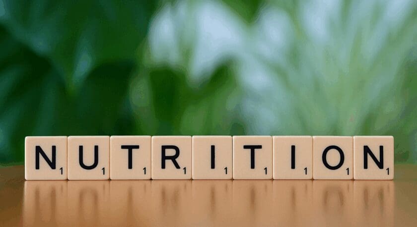 The image features wooden letter tiles arranged to spell the word "NUTRITION." The tiles are set against a blurred green background, suggesting a natural or healthy theme. The surface beneath the tiles is smooth and reflective, enhancing the visual appeal of the arrangement.