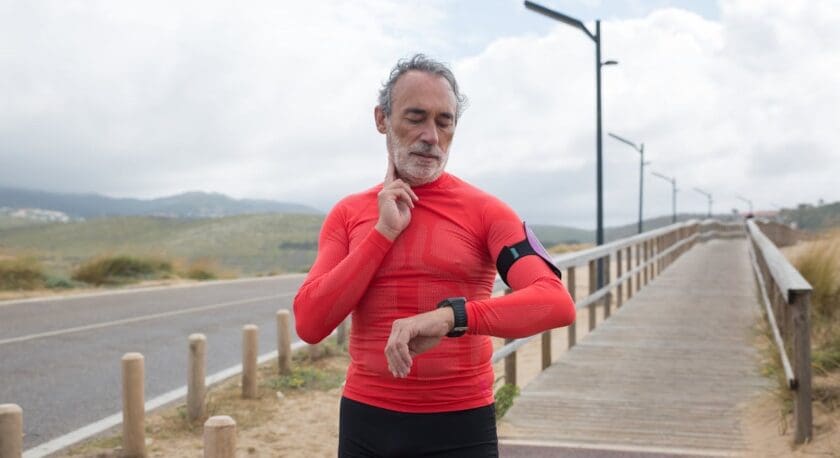 A man in a bright red long-sleeve athletic shirt is checking his smartwatch while standing on a pathway. He has gray hair and a beard, and he appears to be in a thoughtful pose. The background features a wooden boardwalk alongside a road, with green hills and cloudy skies in the distance. The setting suggests an outdoor exercise environment.