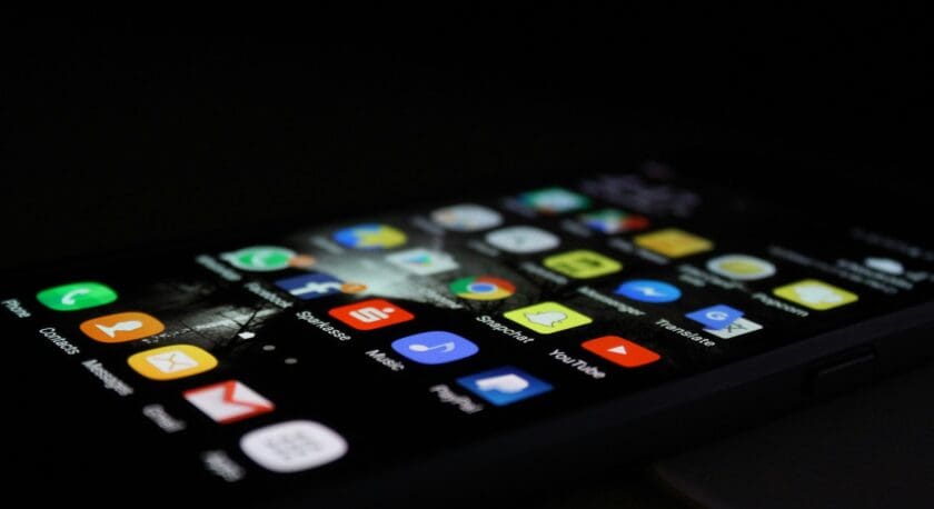 A smartphone is displayed in a dimly lit environment, showcasing its home screen filled with various app icons. The screen features colorful icons for applications such as messaging, social media, music, and utilities. The phone is angled slightly, revealing a dark background and a hint of a surface beneath it. The overall ambiance is modern and tech-focused.