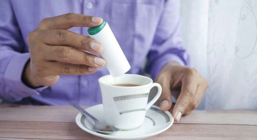 A hand is holding a white container with a green lid above a white cup filled with brown liquid, likely tea or coffee. The cup is placed on a matching saucer, and a silver spoon rests on the saucer. The person is wearing a purple shirt, and there is a sheer curtain in the background, allowing soft light to filter through. The setting appears cozy and inviting.