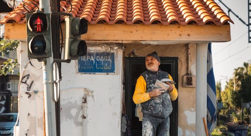 A construction worker stands in the doorway of a small building, wearing a yellow long-sleeve shirt and a gray vest, with paint splatters on his clothes. He holds a trowel and appears to be speaking or gesturing. The building has a terracotta-tiled roof and a weathered white exterior, with a blue street sign that reads "ΠΕΡΑ ΟΔΟΣ." In the background, a traffic light shows a red pedestrian signal, and there are trees and a glimpse of vehicles on the road.