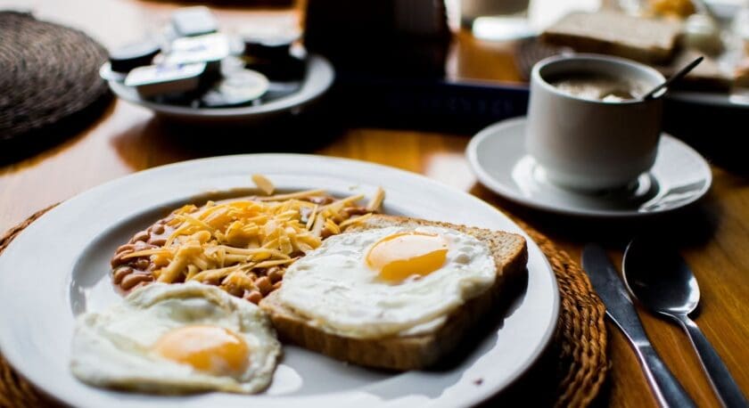 A plate features two sunny-side-up eggs, one resting on a slice of toasted bread. Next to the eggs is a serving of baked beans topped with shredded cheese. The setting includes a cup of coffee and a small plate with condiments in the background, all arranged on a wooden table.