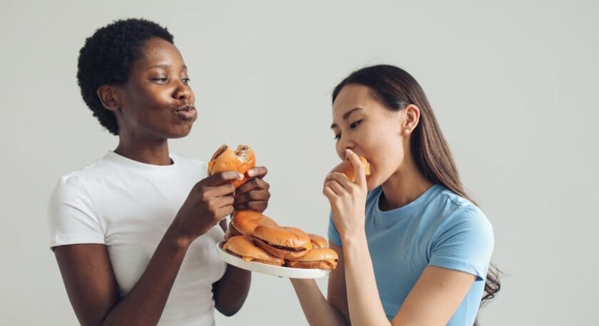 Two women are enjoying burgers in a bright, minimalistic setting. One woman, wearing a white t-shirt, holds two burgers and is playfully pouting. The other woman, dressed in a light blue t-shirt, is taking a bite from a burger while holding a plate stacked with more burgers. The background is a soft, neutral color, enhancing the focus on their expressions and the food.