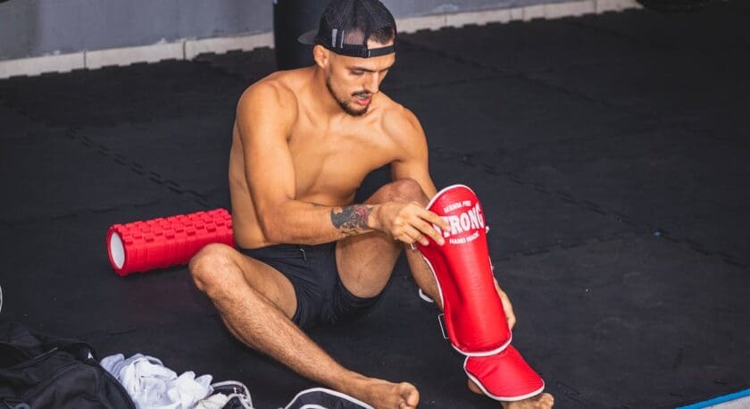 A shirtless man is sitting on a black mat in a training area, focused on putting on a red shin guard. He has short hair and is wearing a black cap and black shorts. Nearby, a red foam roller and a black gym bag are visible, along with some white clothing and black training shoes on the floor. The background features a gray wall and a punching bag.