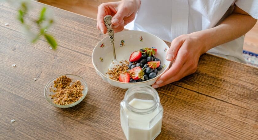 A person is holding a decorative bowl filled with fresh blueberries and sliced strawberries, with a sprinkle of granola on top. A vintage-style spoon is in the bowl. Next to it, there is a small glass dish containing more granola. A jar of milk is placed on the wooden table, completing the breakfast scene. The background features a soft, blurred interior setting.