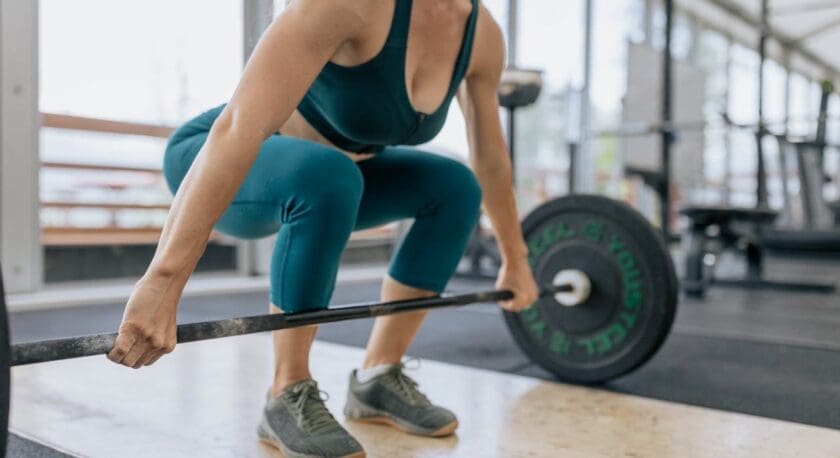 A person is performing a deadlift in a gym setting. They are wearing a teal sports bra and matching leggings, with gray athletic shoes. The individual is crouched down, gripping a barbell loaded with weights. The gym has large windows, allowing natural light to illuminate the space, and various gym equipment is visible in the background. The flooring is a combination of wooden and rubber surfaces.