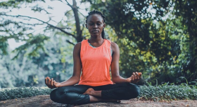 A woman is meditating outdoors, seated cross-legged on a rock. She wears a bright orange tank top and black leggings. Her eyes are closed, and her hands rest on her knees in a mudra position. The background features lush greenery and trees, creating a serene and peaceful atmosphere.