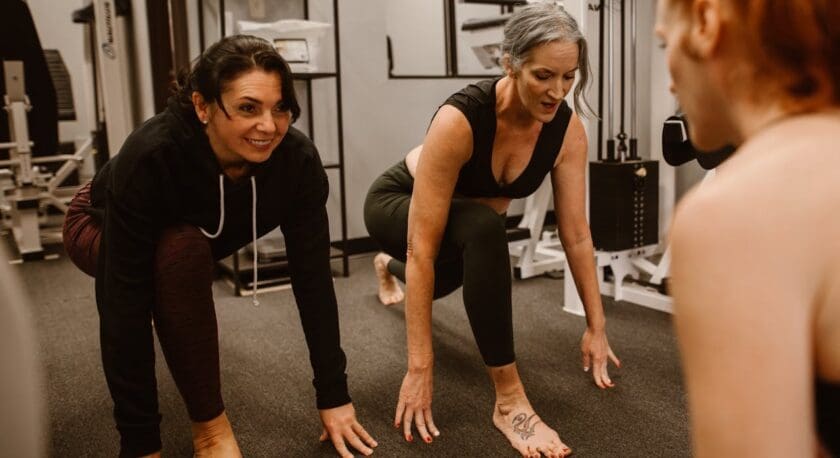 Two women are engaged in a workout session in a gym. One woman, wearing a black hoodie and maroon leggings, is smiling and positioned in a crouched stance. The other woman, dressed in a black crop top and dark green leggings, is also in a crouched position, focusing intently. Both are barefoot and appear to be demonstrating or practicing exercises. The gym has various fitness equipment in the background, and the flooring is dark.