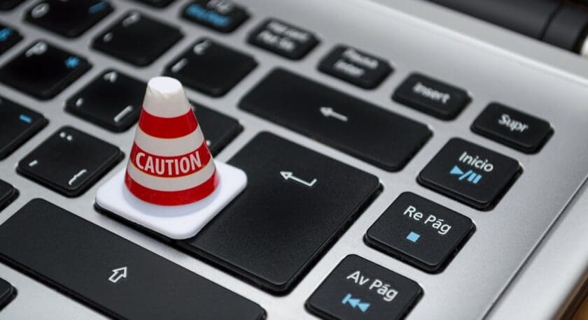 A small red and white striped caution cone is placed on a laptop keyboard, specifically on the arrow key. The keyboard features black keys with white and blue lettering, including keys labeled "Inicio," "Re Pág," and "Av Pág." The laptop's surface is silver, and the image is taken from a close-up angle, focusing on the cone and the surrounding keys.