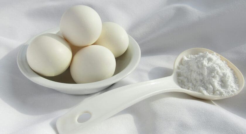 A small white bowl holds four white eggs, arranged neatly. Next to the bowl, a white measuring spoon filled with a fine white powder rests on a smooth, white fabric surface. The overall composition is simple and clean, emphasizing the eggs and the powder.
