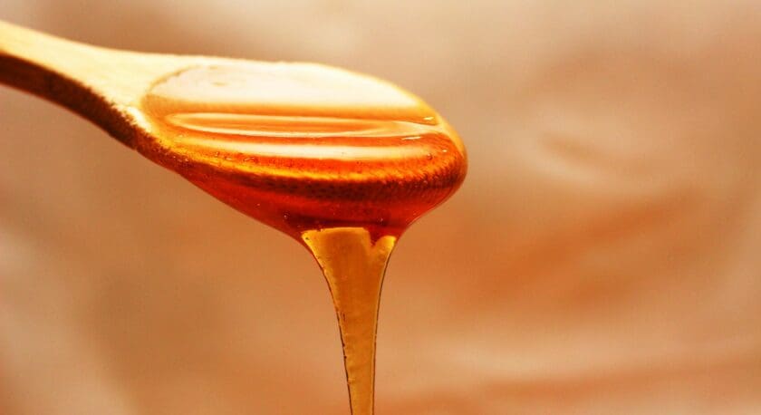 A wooden spoon is shown with a glossy, amber-colored honey dripping from its tip. The honey has a smooth texture and is glistening under soft lighting, creating a warm and inviting appearance. The background is softly blurred, emphasizing the spoon and the honey.