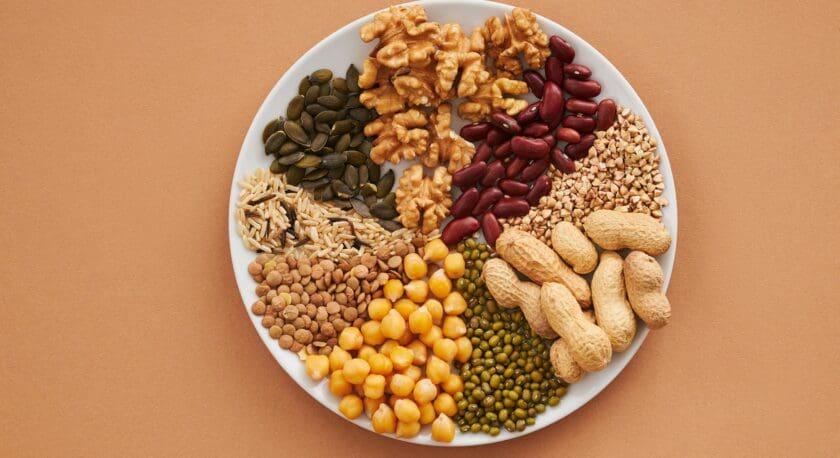 A round white plate is filled with a variety of nuts and legumes, arranged in sections. The top left features dark green pumpkin seeds, followed by a cluster of walnut halves. Next, there are red kidney beans and a mix of small grains, including buckwheat. The bottom left showcases lentils and yellow chickpeas, while green mung beans are positioned next to them. Finally, the plate includes several whole peanuts in their shells. The background is a warm, light brown color.