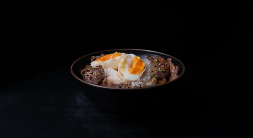 A black bowl filled with a serving of rice topped with pieces of beef and two soft-boiled eggs. The eggs have bright orange yolks that are slightly runny. The dish is garnished with green onions and sesame seeds, set against a dark background that enhances the colors and textures of the food.