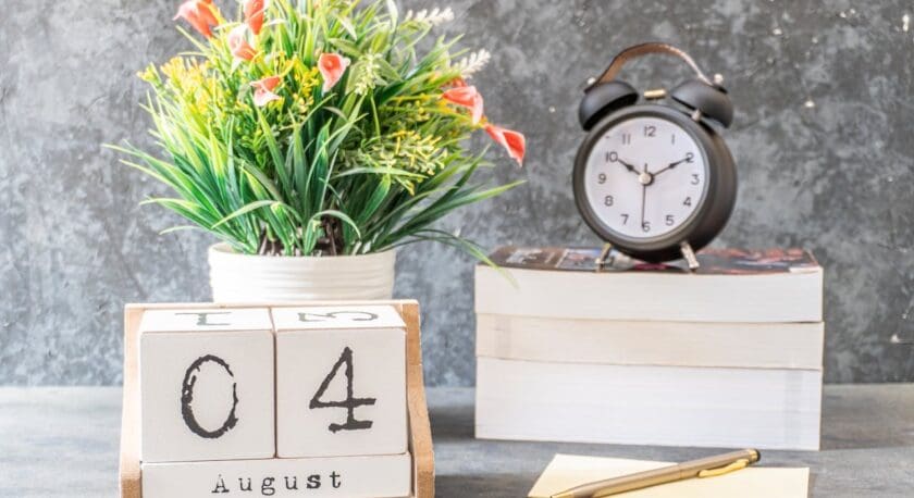 A decorative arrangement features a block calendar displaying "04" and "August" on a wooden stand. Beside it, a black alarm clock shows the time, with a white face and black numbers. In the background, a potted plant with vibrant green leaves and orange flowers adds a touch of color. There are also a few stacked books and a gold pen resting on a yellow notepad. The overall setting has a modern and organized aesthetic.