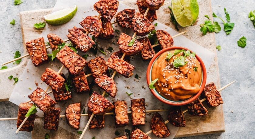 Skewers of grilled tempeh are arranged on a wooden board, garnished with sesame seeds and fresh cilantro. A small bowl of creamy peanut sauce sits nearby, topped with additional cilantro and sesame seeds. Lime wedges are placed alongside the skewers, adding a pop of color. The background features a light gray surface, enhancing the vibrant presentation of the dish.