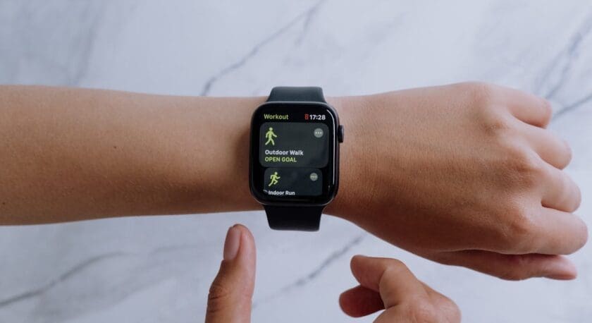 A close-up of a wrist wearing a black smartwatch displaying workout options. The screen shows "Outdoor Walk" with an "OPEN GOAL" option and "Indoor Run" below it. A finger is poised to interact with the watch, while the background features a light marble texture.