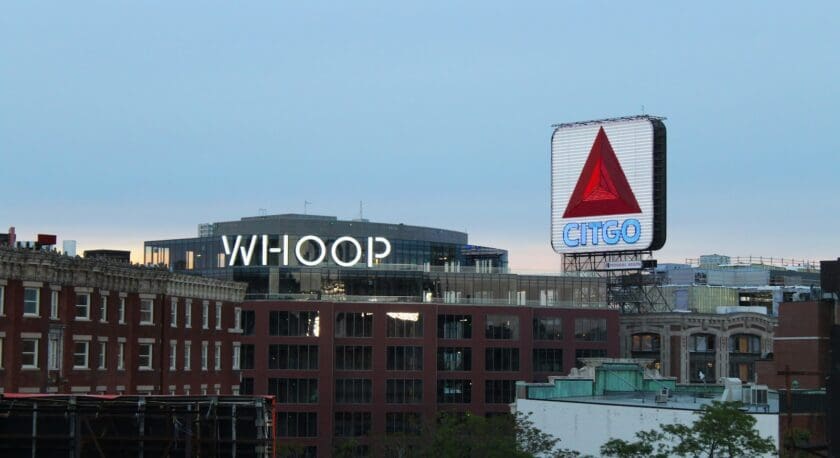 A cityscape features two prominent signs. On the left, a building displays the word "WHOOP" in large, illuminated letters. To the right, a tall, illuminated sign features the CITGO logo, which includes a red triangular symbol and the brand name in blue. The background shows a mix of modern and historic architecture, with a cloudy sky above.