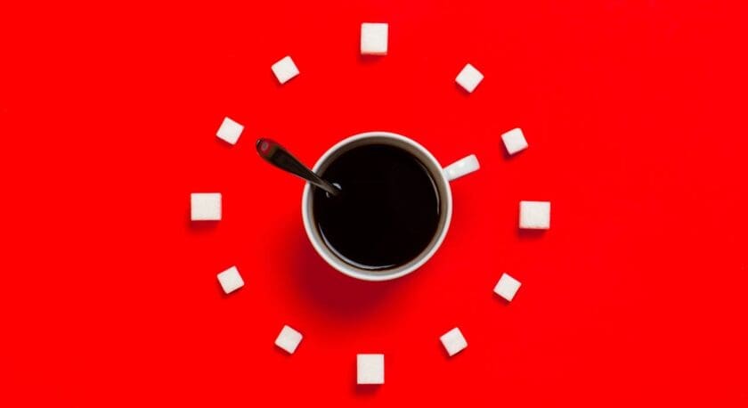 A white cup filled with black coffee sits at the center of a bright red surface. Surrounding the cup are several small, white sugar cubes arranged in a circular pattern. A silver spoon rests inside the cup, partially submerged in the coffee. The overall composition creates a striking contrast between the colors and shapes.