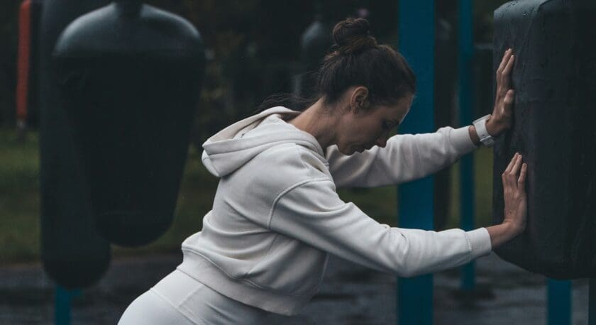 A woman in a white hoodie and white leggings is leaning forward with both hands pressing against a black padded surface, appearing to stretch or rest. She has her hair tied up in a bun and is wearing a white watch. The background shows outdoor gym equipment and a dim, overcast setting.